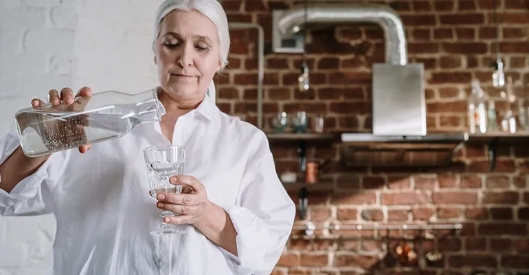 Kvinna klädd i vitt står i ett kök och häller upp i vatten från en karaff i ett glas 